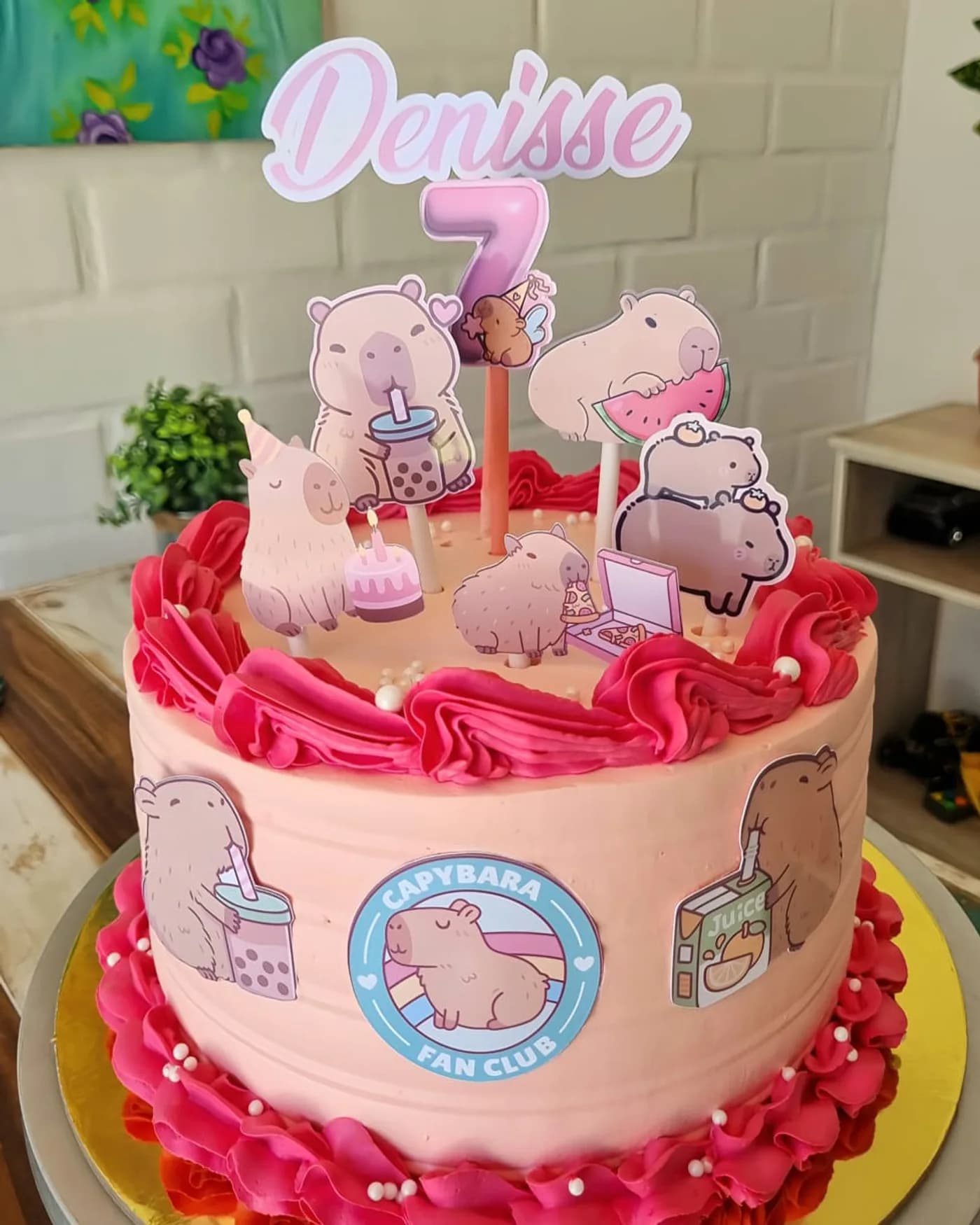 Torta rosada con pétalos de crema fucsia y toppers de capibaras con el nombre Denisse